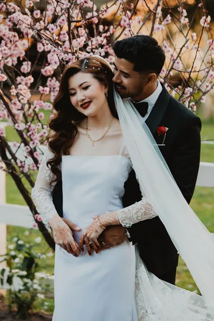 The bride smiling beneath the spring blossom, her groom behind her